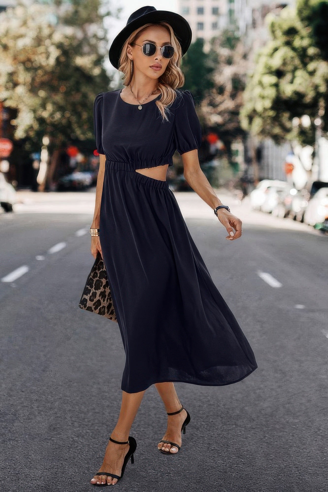 Woman in navy midi dress with side cutout, black hat, sunglasses, and leopard clutch walking outdoors