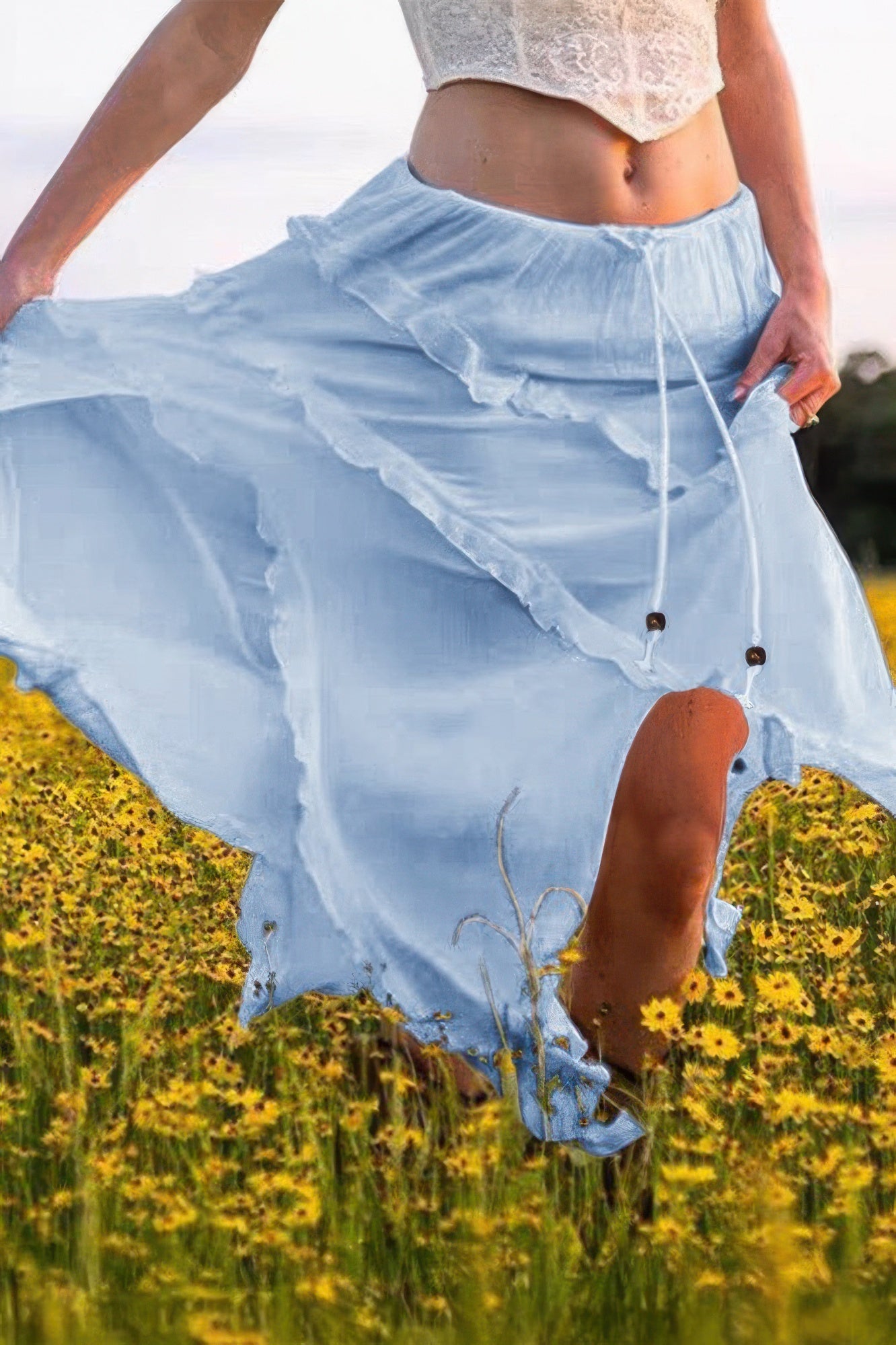 Woman in flowing light blue skirt with lace top, standing in yellow wildflower field, stylish summer outfit