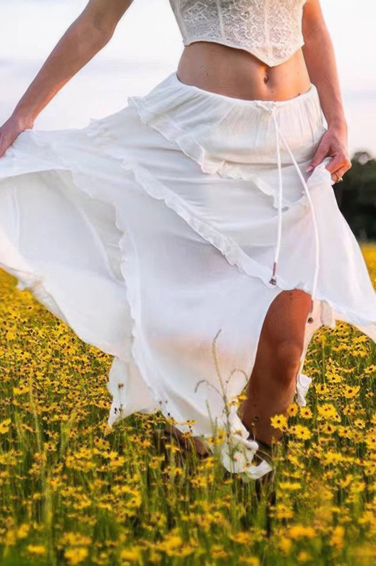 Woman in flowing white skirt and lace top twirling in yellow flower field, stylish summer outfit