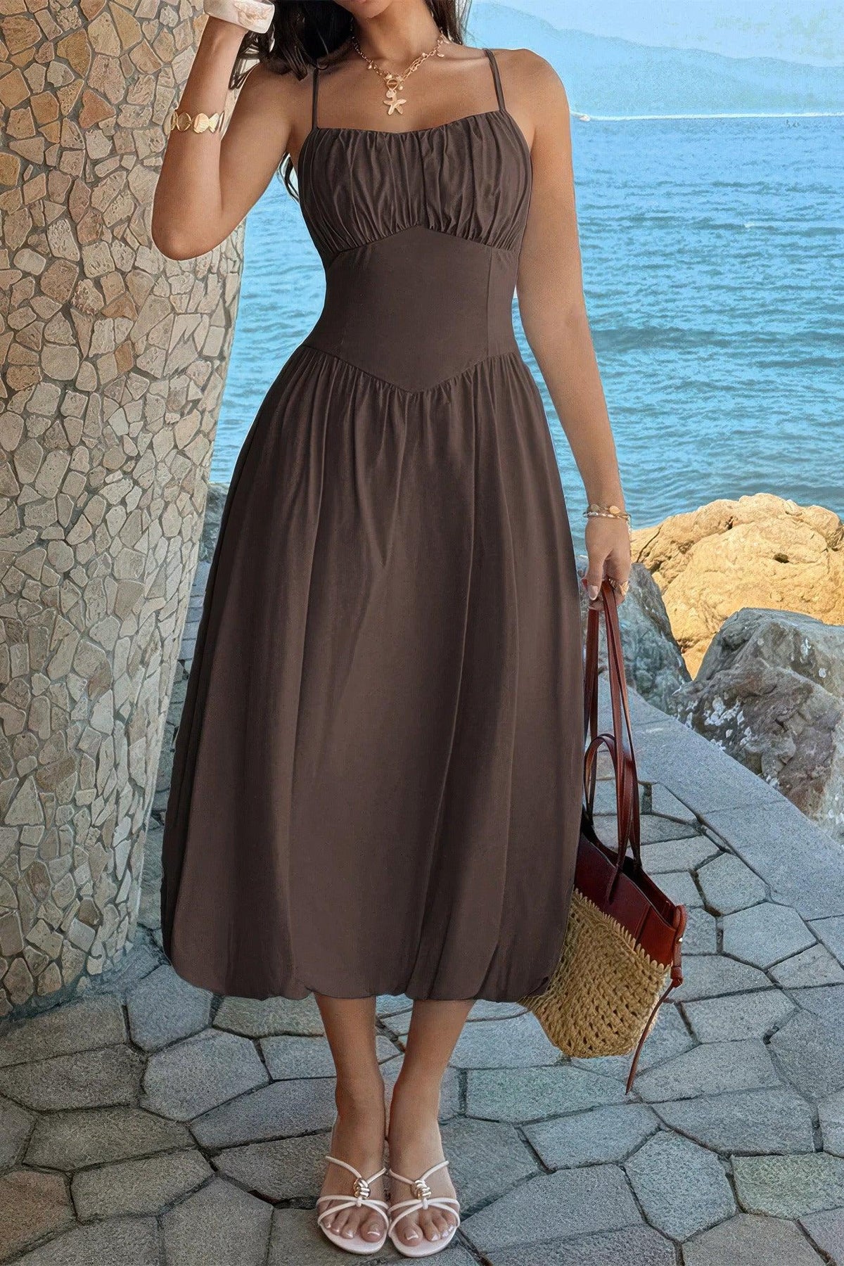 Brown midi dress with ruched bodice, thin straps, and flowy skirt, styled with sandals and straw bag by the sea