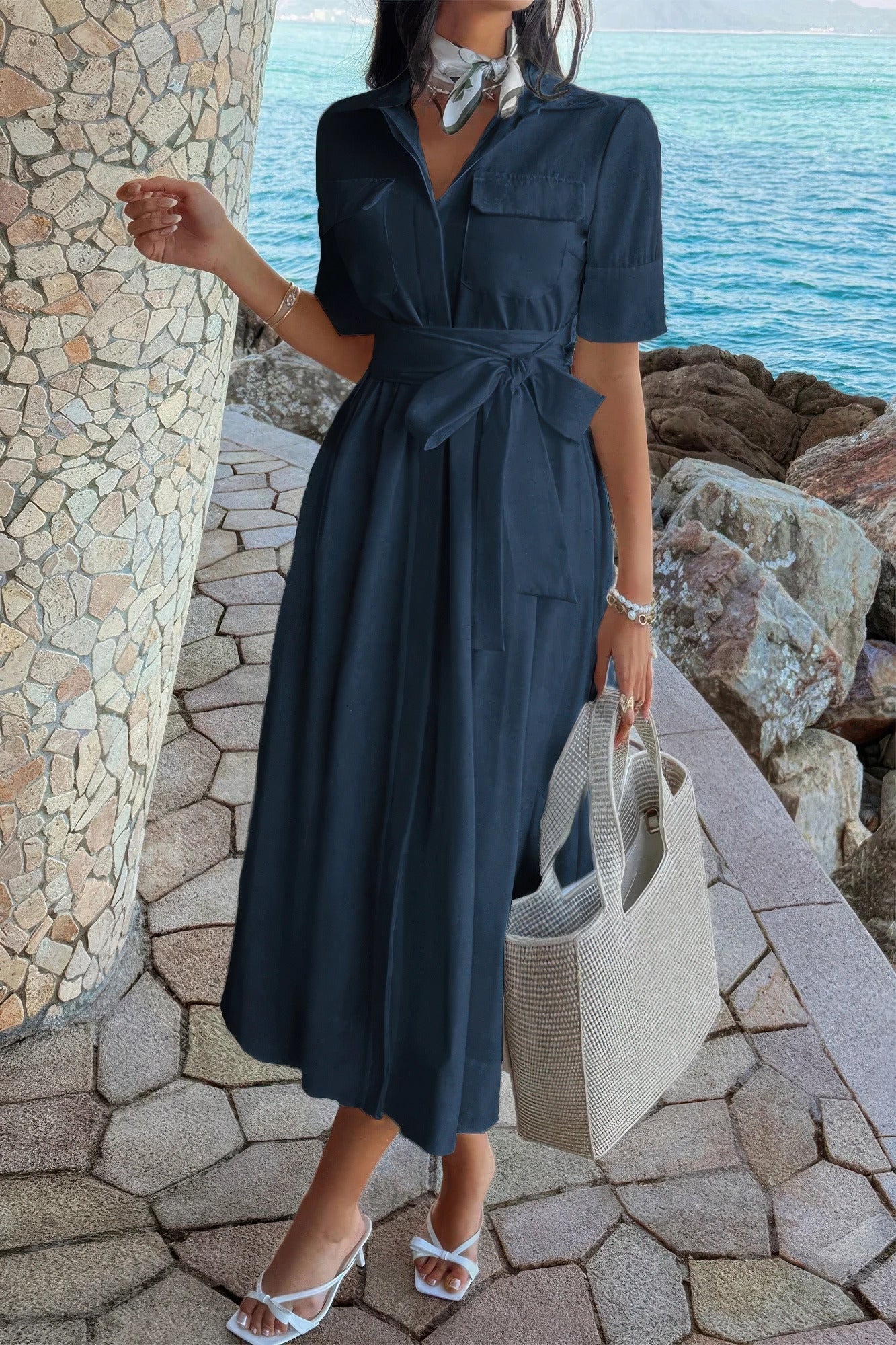 Navy blue midi dress with tie waist, white sandals, and woven tote bag by the seaside
