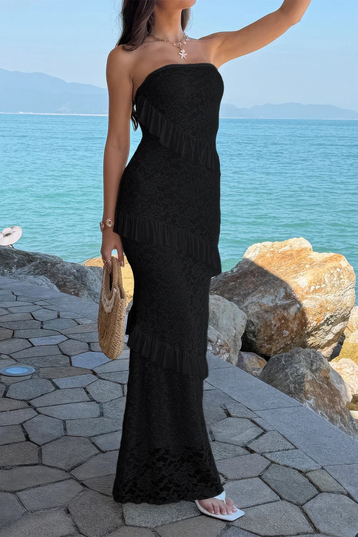 Black strapless ruffle dress with floral lace, worn by woman at seaside, holding woven bag.